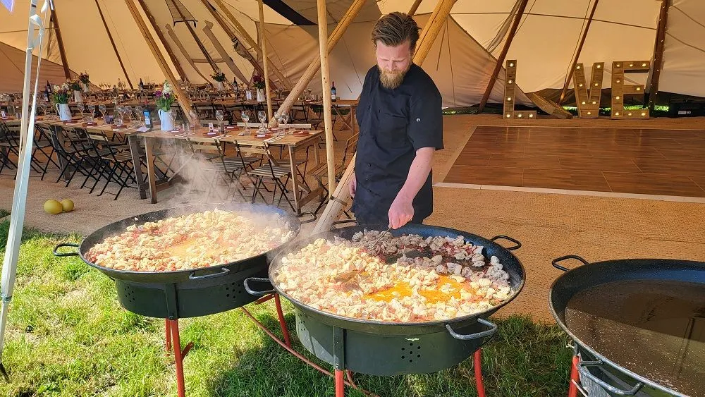 A chef live-cooking paella in large pans at a dressed wedding tipi venue